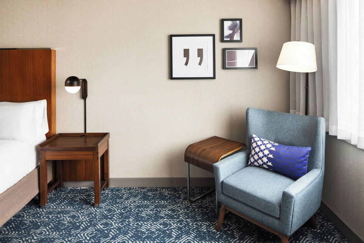 A hotel room with a bed, a chair, a nightstand, a lamp, and a wall with framed pictures.