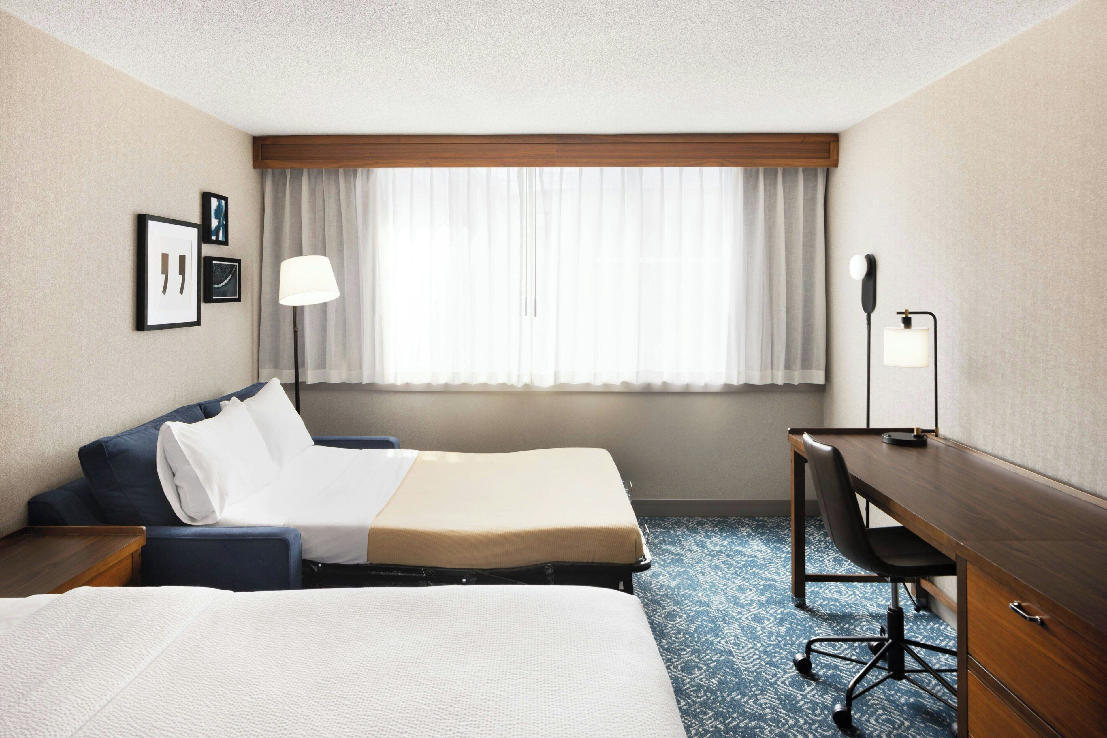 Una habitación de hotel con dos camas, un escritorio, una silla, una lámpara y una ventana con cortinas.