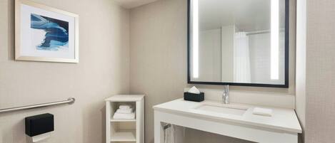 A modern bathroom with a white vanity, a large mirror, a framed artwork, a towel rack, and a hand sanitizer dispenser.