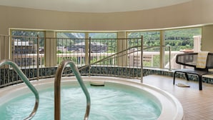 Un bain à remous doté de barrières en métal, une vue sur les bâtiments et les montagnes à travers de grandes fenêtres, et un fauteuil avec un coussin à rayures.