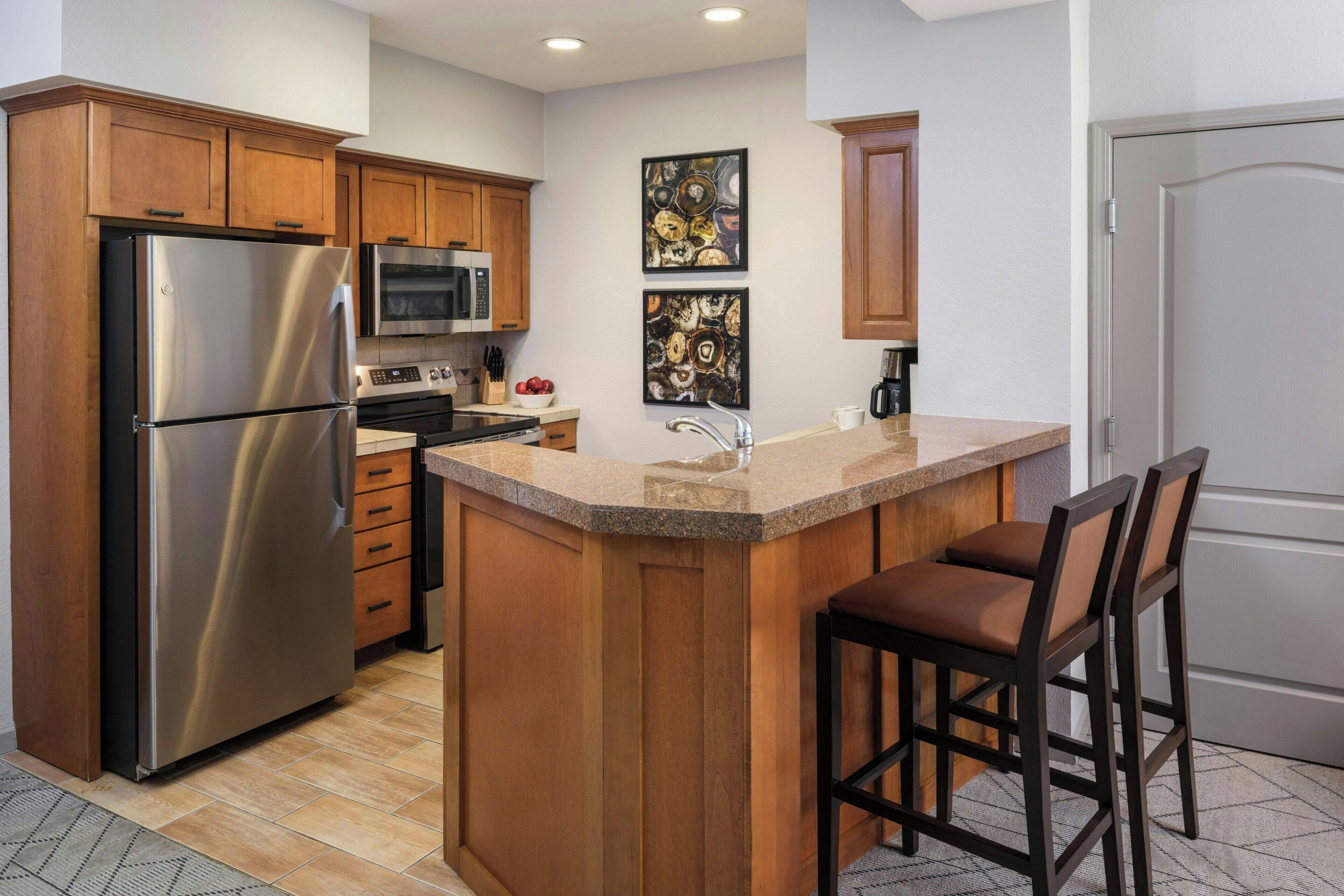 A kitchen with wooden cabinets, stainless steel appliances, a granite countertop, and two bar stools.