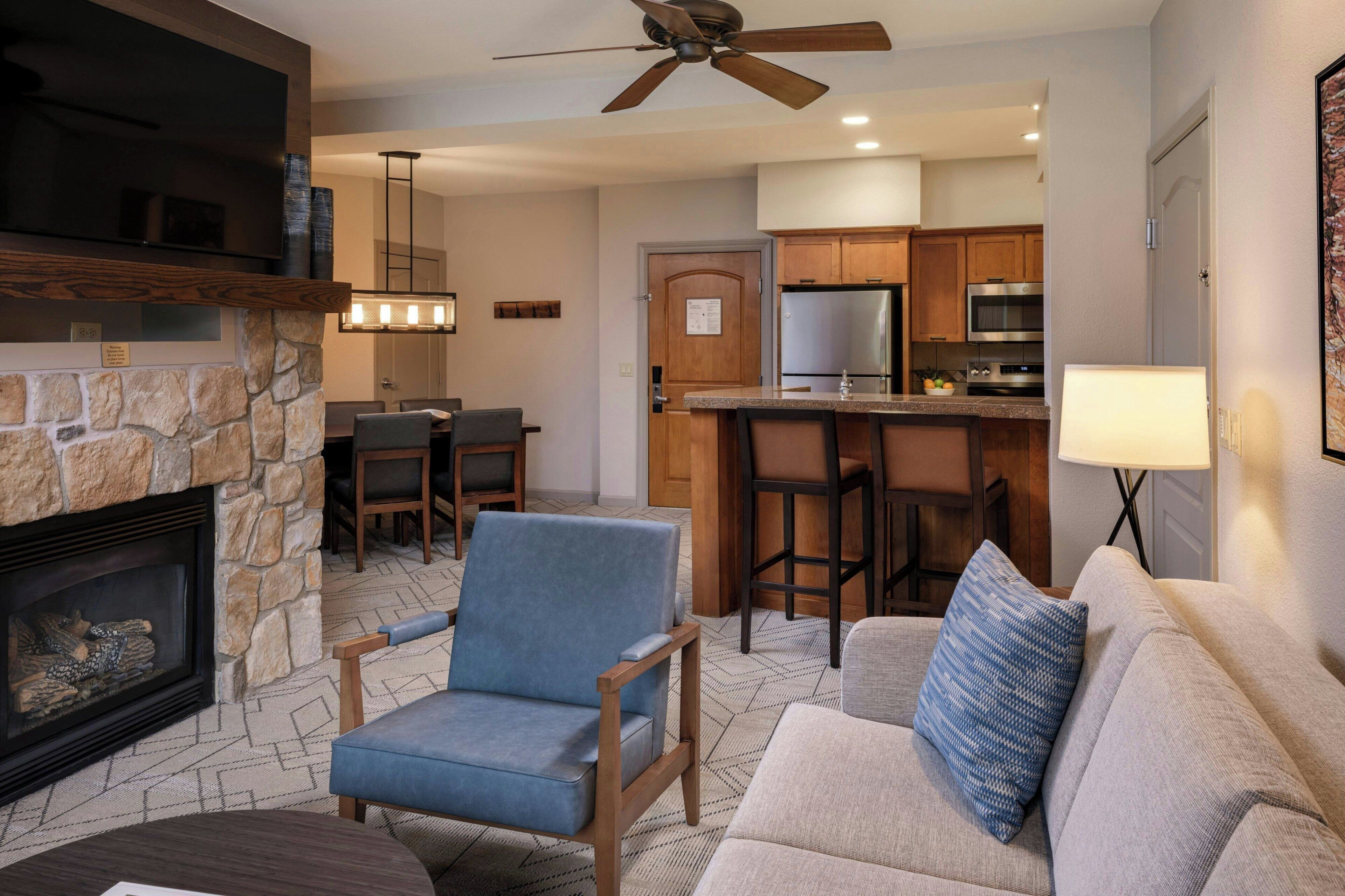 A living room with a stone fireplace, a blue armchair, a gray sofa, and a ceiling fan.