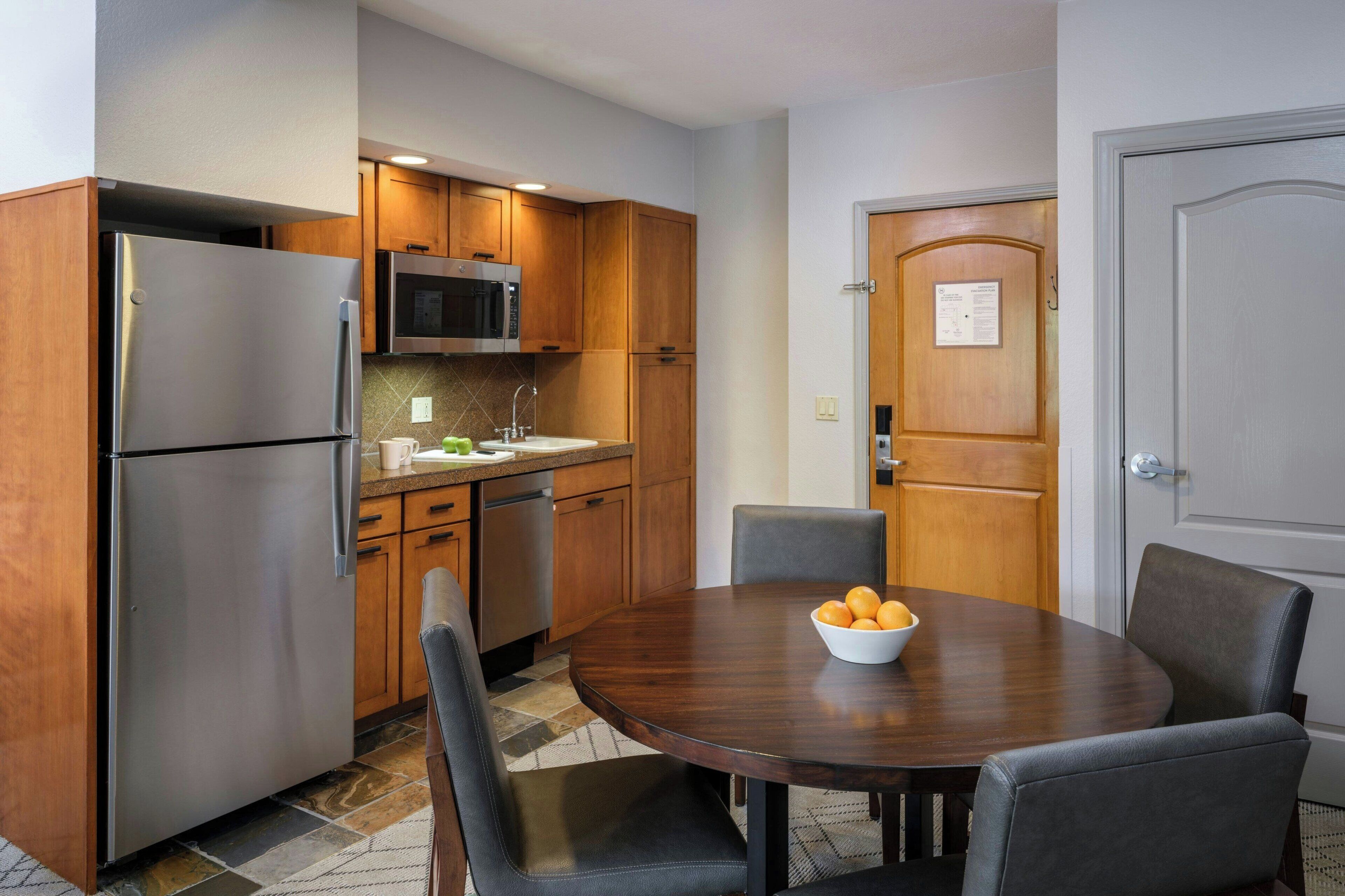 Una cocina compacta con refrigerador, microondas y gabinetes de madera. Cuenta con una mesa de comedor con sillas y un tazón de fruta.