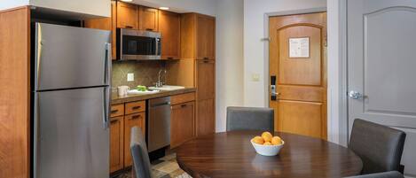 Una cocina compacta con refrigerador, microondas y gabinetes de madera. Cuenta con una mesa de comedor con sillas y un tazón de fruta.