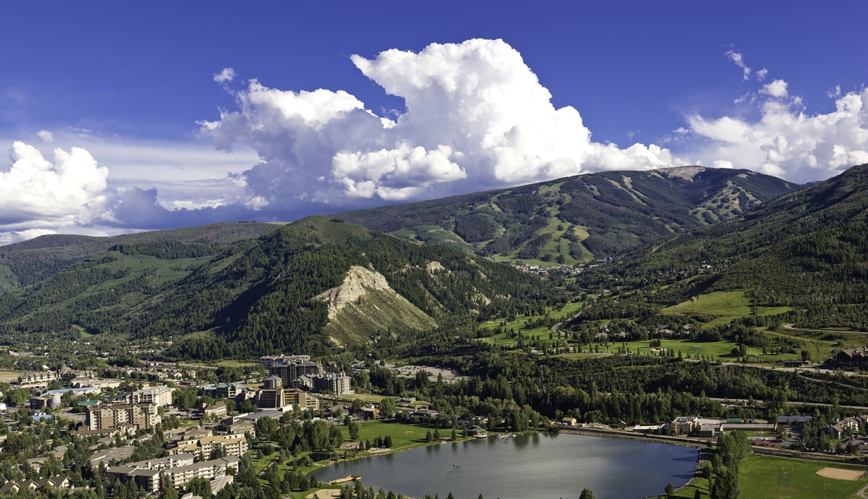 Paisagem montanhosa com um lago, uma cidade com edifícios e céu claro.
