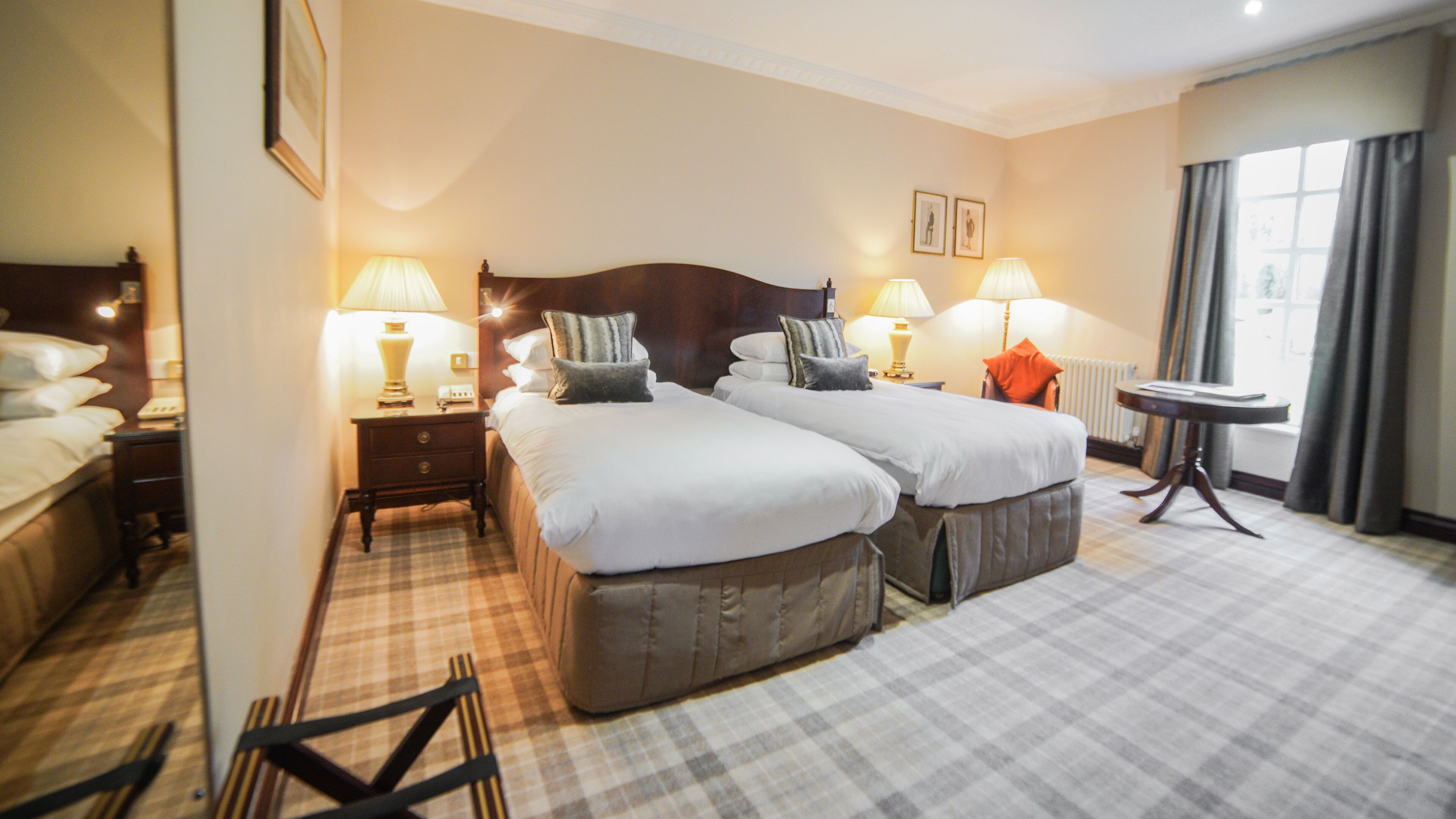 A hotel room with two beds, a small table, a chair, a lamp, and a window with curtains.