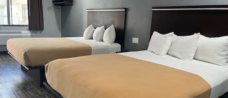 A hotel room with two beds, each with a headboard and pillows. There is a window with curtains, an air conditioning unit, and a wall-mounted shelf.