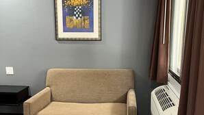 A living room with a beige sofa, a framed artwork on the wall, a window with curtains, and a white radiator. -  Laguna Inn & Suites (San Juan Capistrano)