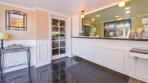 A hotel lobby with a reception desk, a mirror, a door leading to a storage area, and a window with a view of the outside. -  Laguna Inn & Suites (San Juan Capistrano)