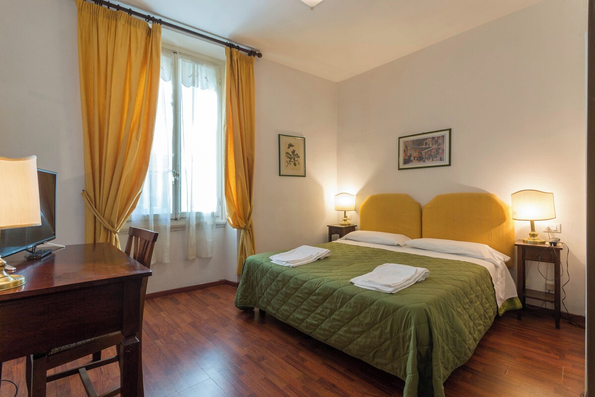 Habitación de hotel con cama, dos lámparas de noche, un escritorio con computadora y una ventana con cortinas.
