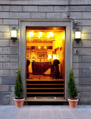 A hotel entrance with a glass door, a reception desk inside, and two potted plants outside. - Hotel Rex (Florence)