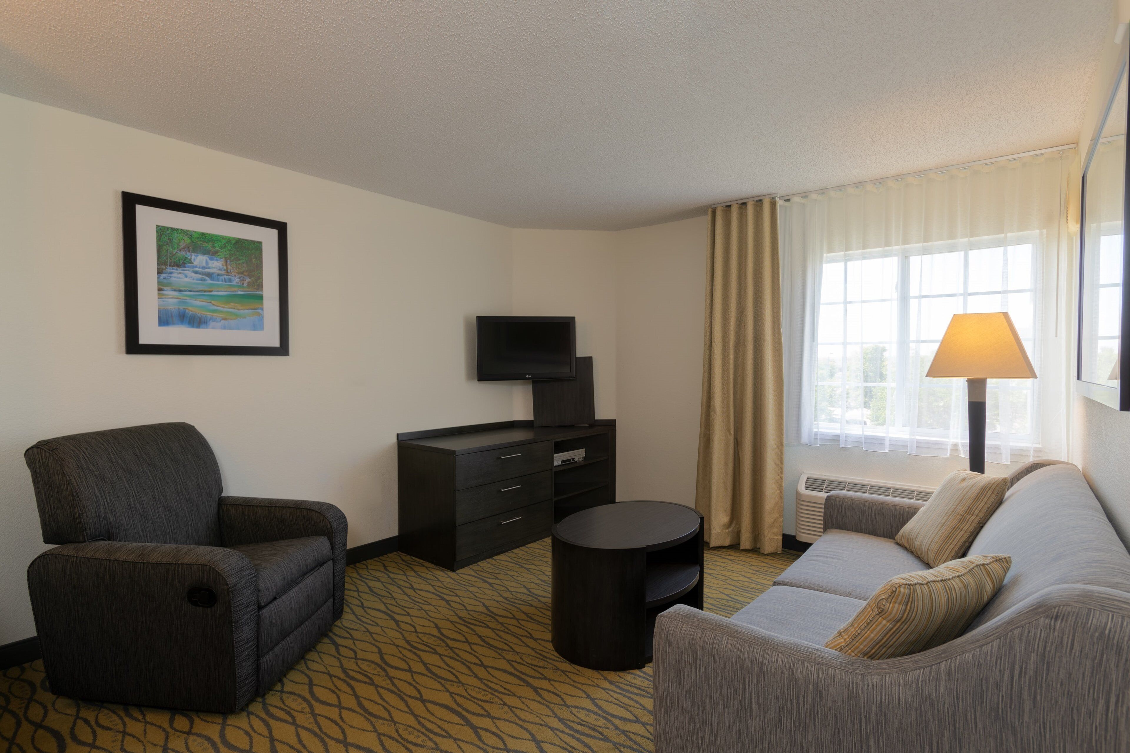 A hotel room with a sofa, armchair, TV stand, and a framed picture on the wall.