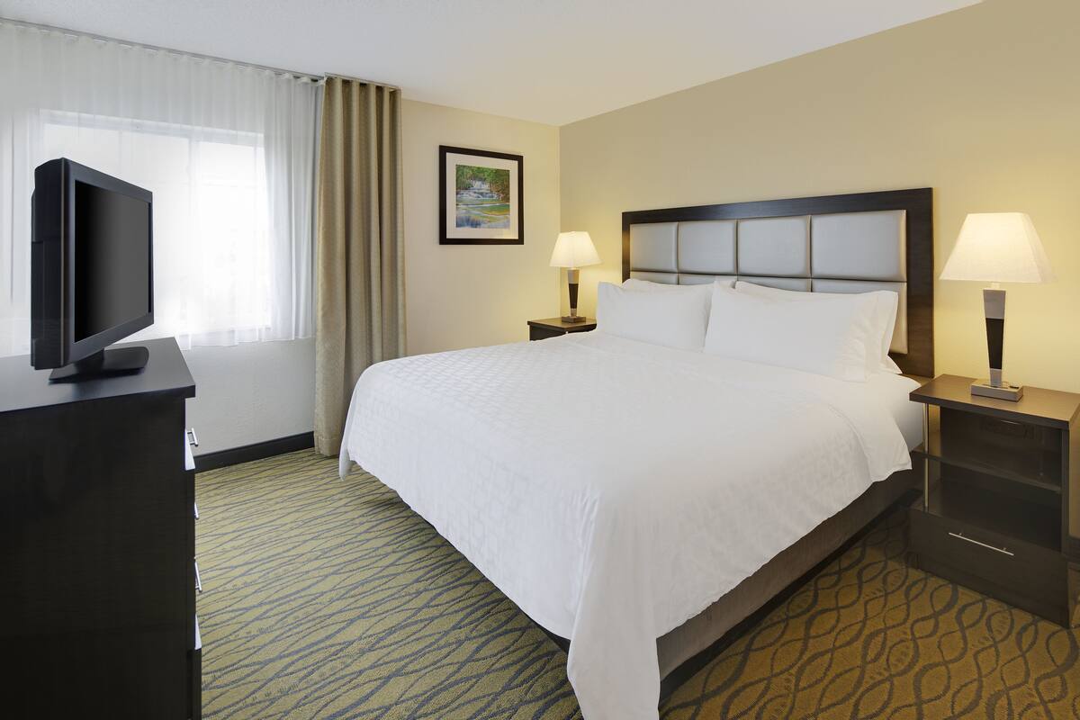A hotel room with a large bed, two bedside tables with lamps, a flat-screen TV on a stand, a framed picture on the wall, and a window with curtains.
