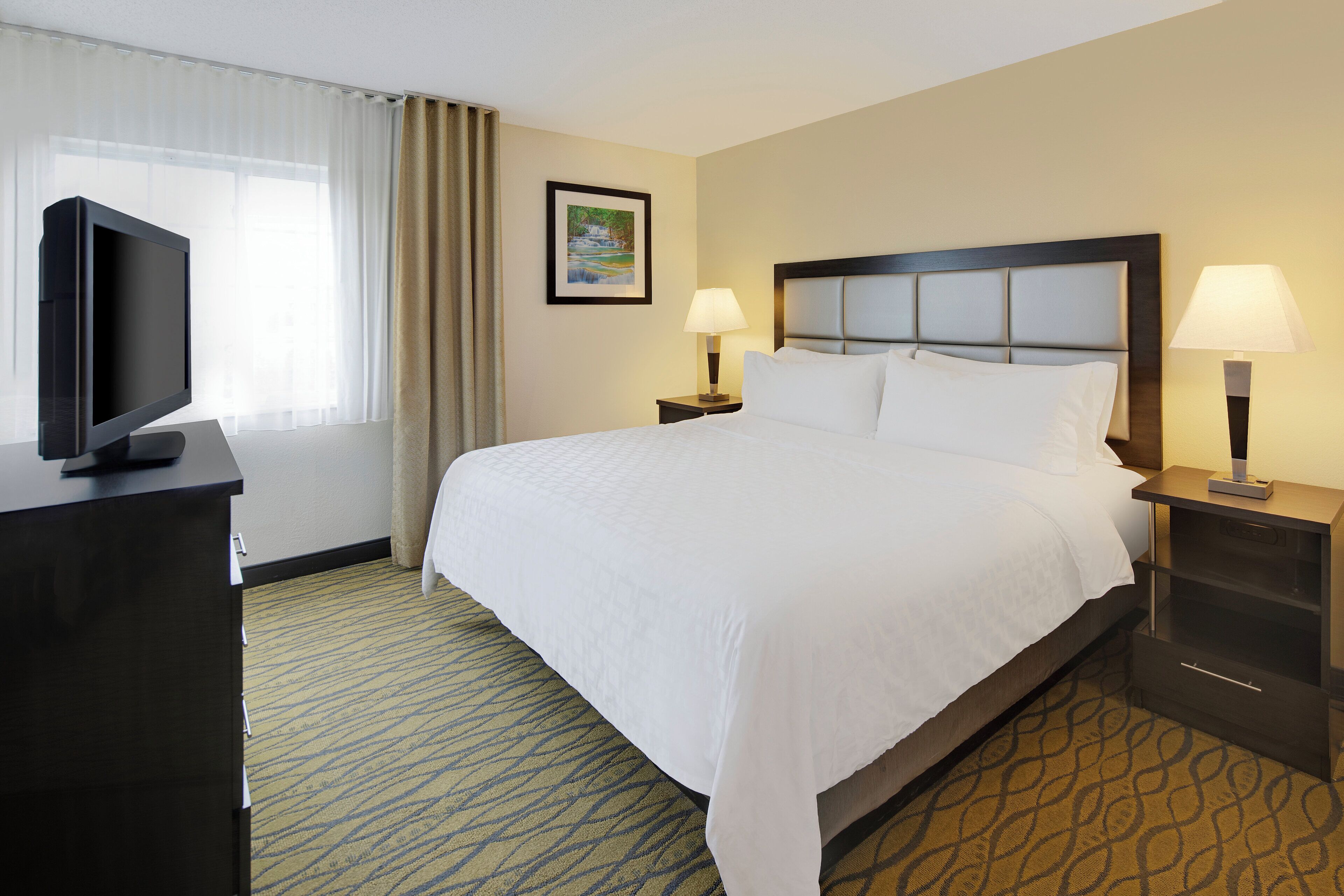A hotel room with a large bed, two bedside lamps, a television on a desk, and a framed picture on the wall.