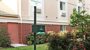 Un bote de basura con un contenedor de reciclaje adjunto, un letrero que dice 'Limpie después de su mascota', y un edificio de ladrillo con ventanas y unidades de aire acondicionado al fondo.