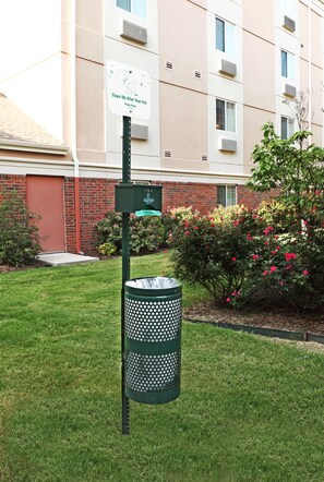 Un bote de basura con un contenedor de reciclaje adjunto, un letrero que dice 'Limpie después de su mascota', y un edificio de ladrillo con ventanas y unidades de aire acondicionado al fondo.