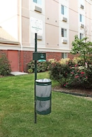 Un bote de basura con un contenedor de reciclaje adjunto, un letrero que dice 'Limpie después de su mascota', y un edificio de ladrillo con ventanas y unidades de aire acondicionado al fondo.