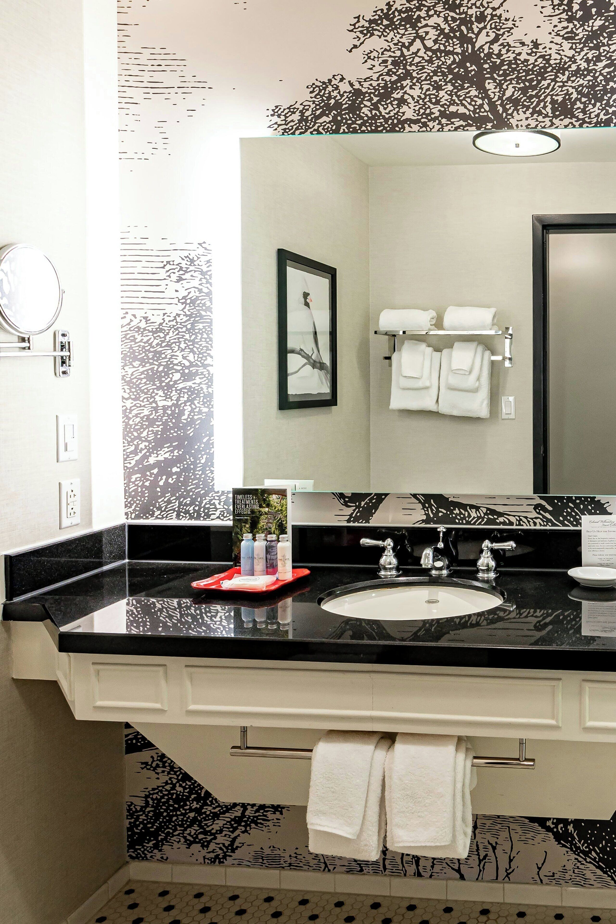 Un baño moderno con encimera negra, lavabo blanco y un espejo grande. Hay toallas en un perchero y un cuadro en la pared.