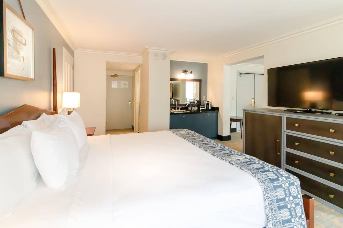 A hotel room with a large bed, a television, a dresser, and a kitchenette in the background.