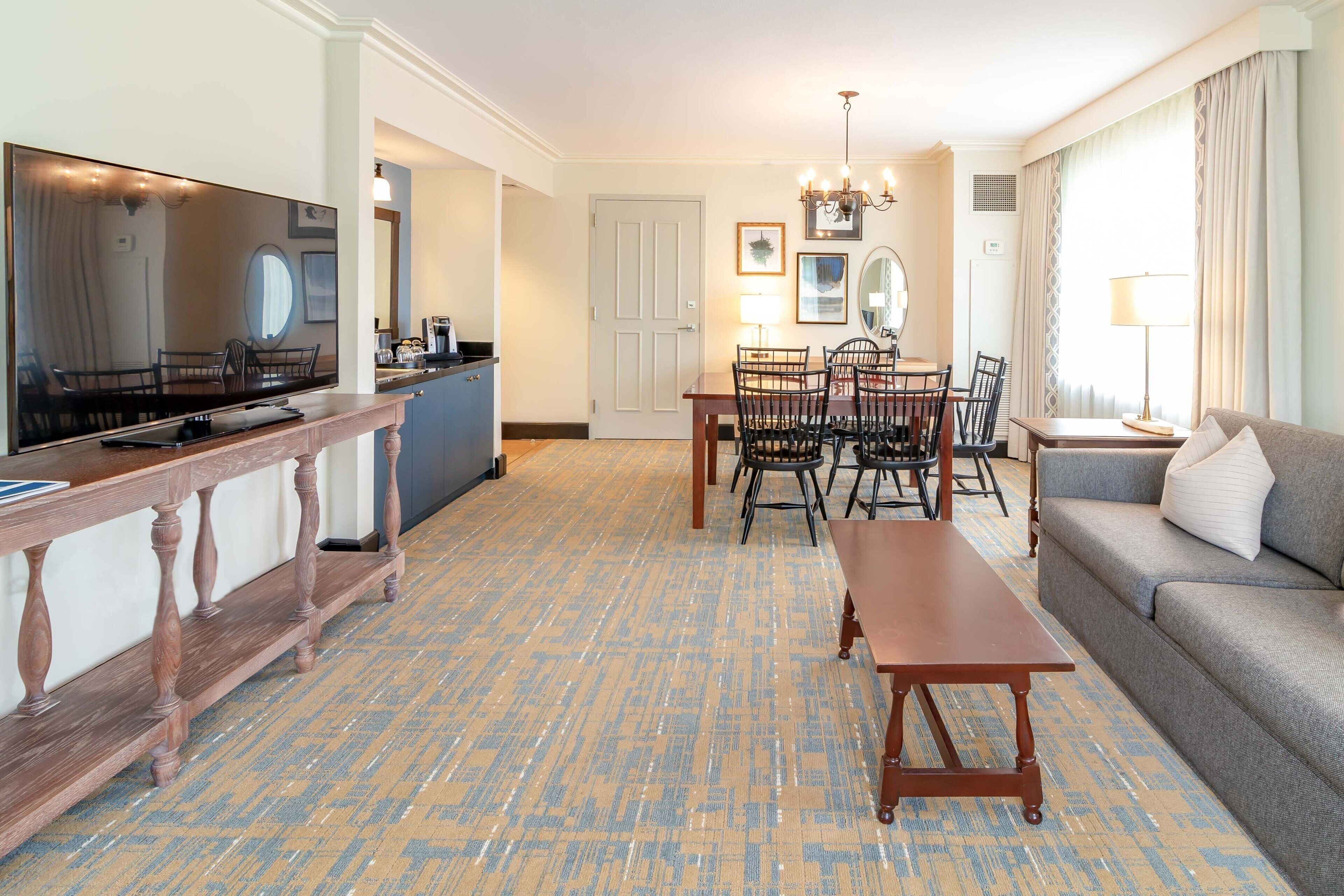 A hotel room with a dining area, a sofa, a TV, and a wooden bench.