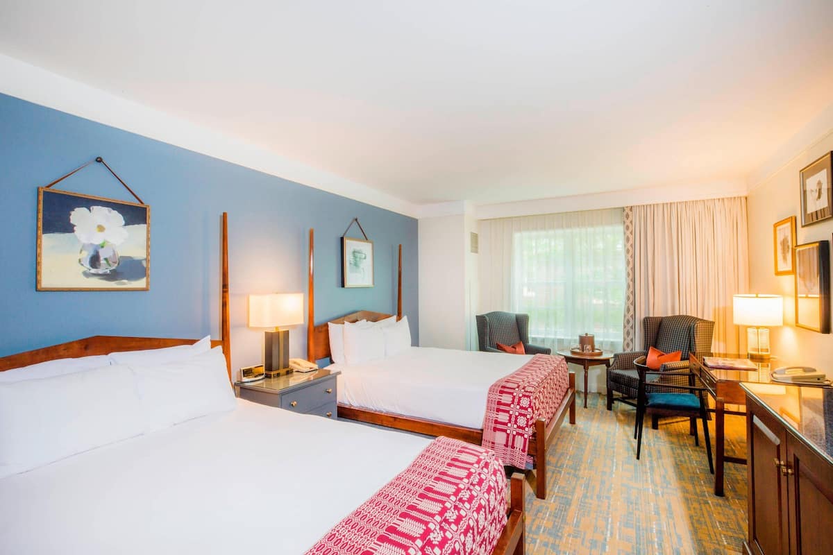 A hotel room with two beds, a desk, a chair, a small table, and a window with curtains.