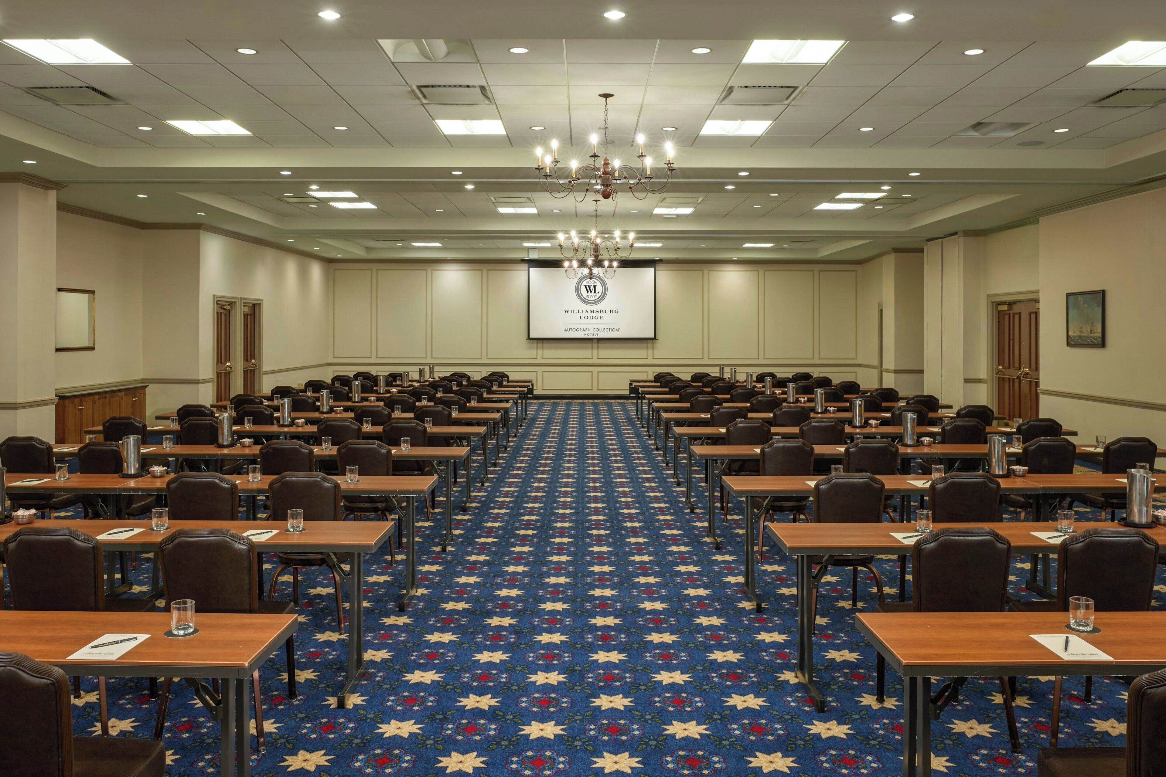 Un salón de conferencias con filas de mesas, una pantalla de proyector y una araña.