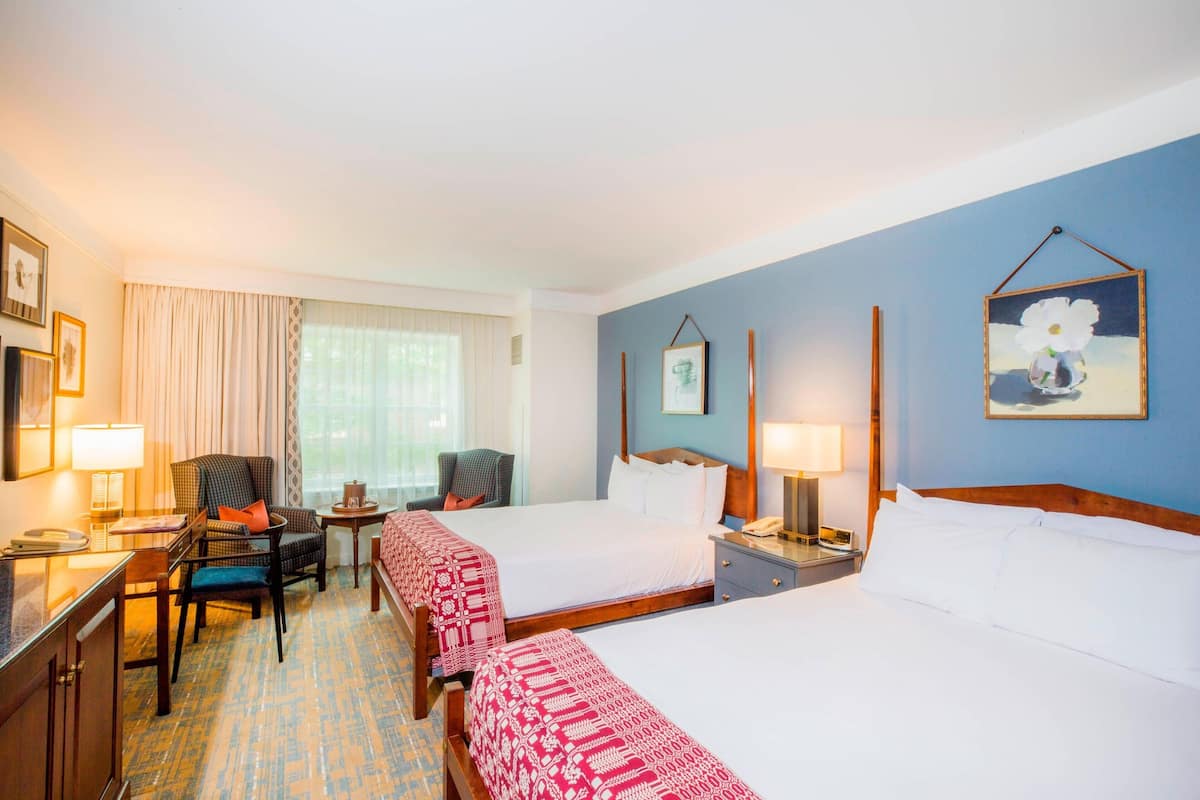 A hotel room with two beds, a desk, a chair, a small table, and a window with curtains.