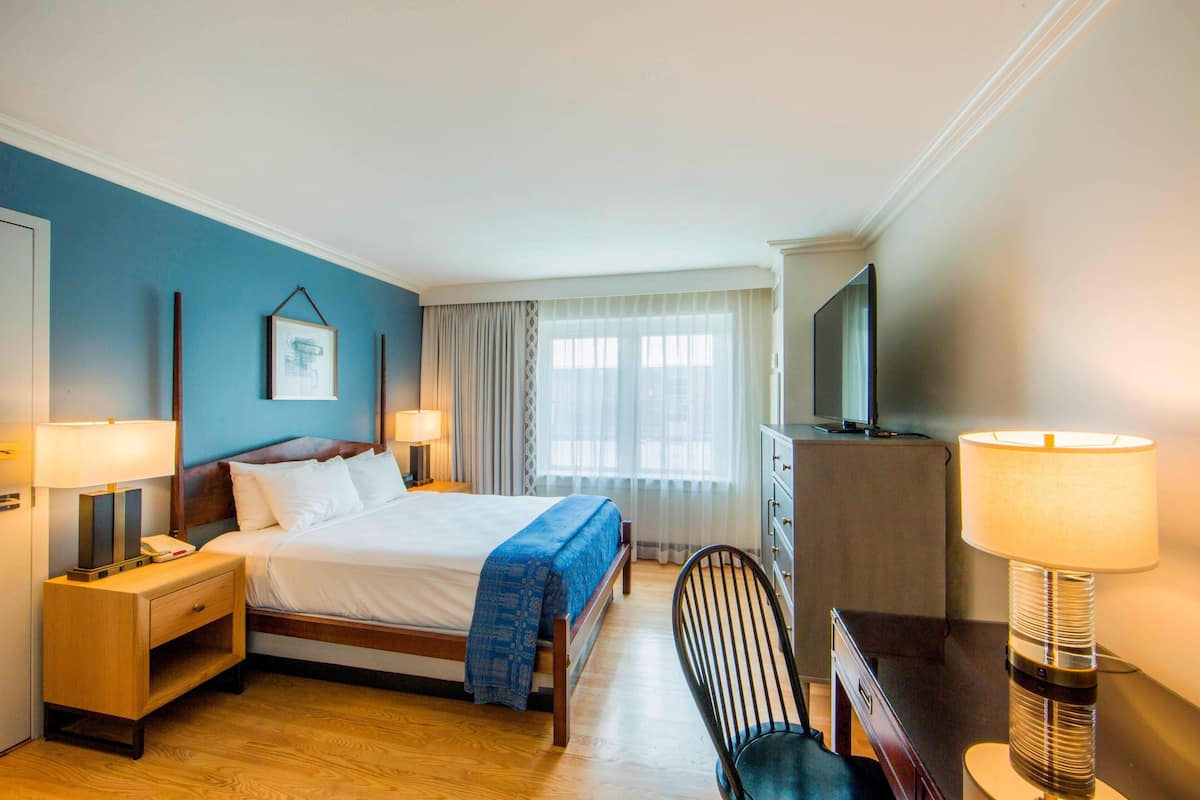 A hotel room with a bed, bedside tables, a desk, a chair, a television, and a window with curtains.