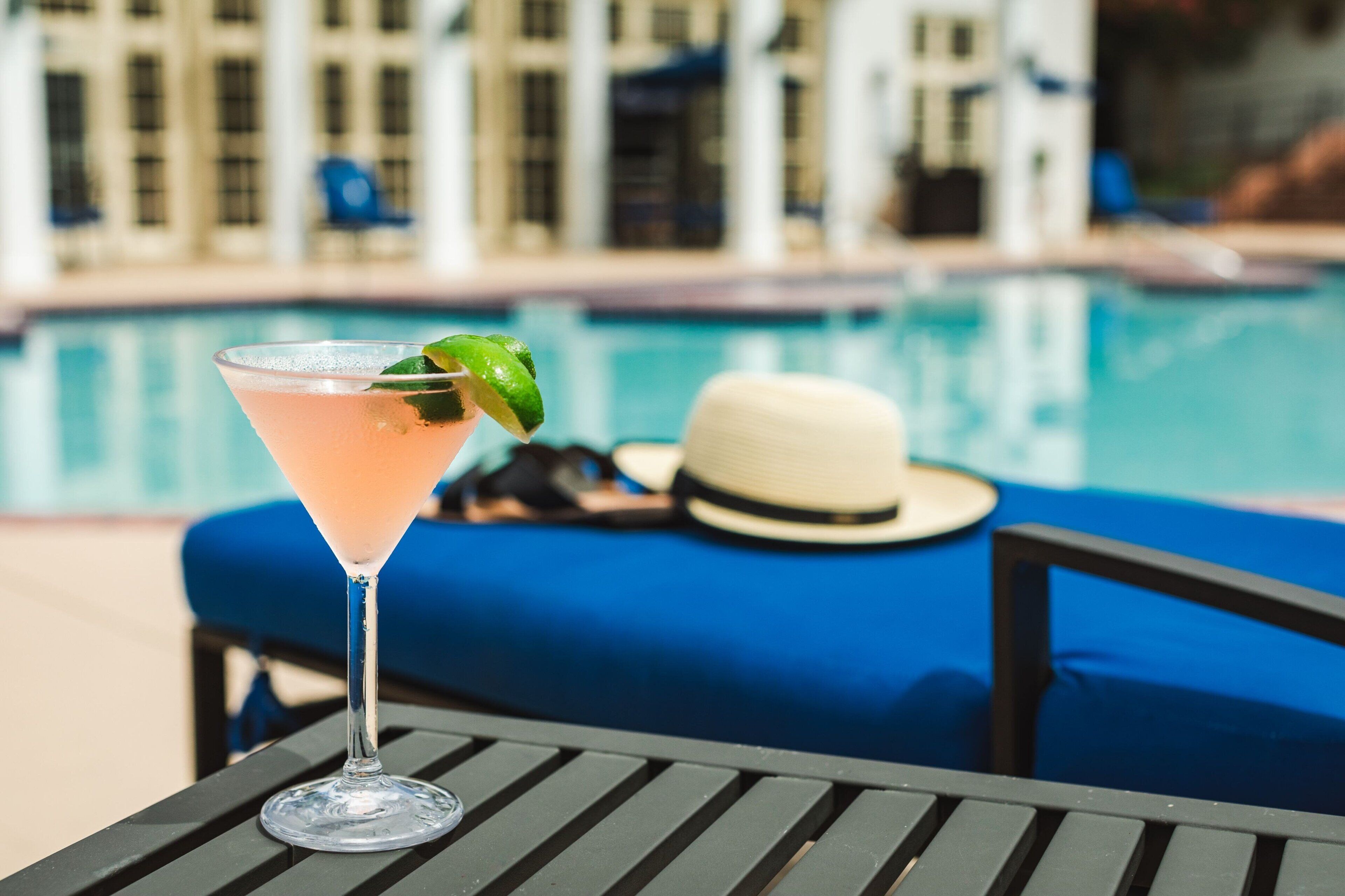 A cocktail on a table near a poolside lounge with a hat and sunglasses on the bench.
