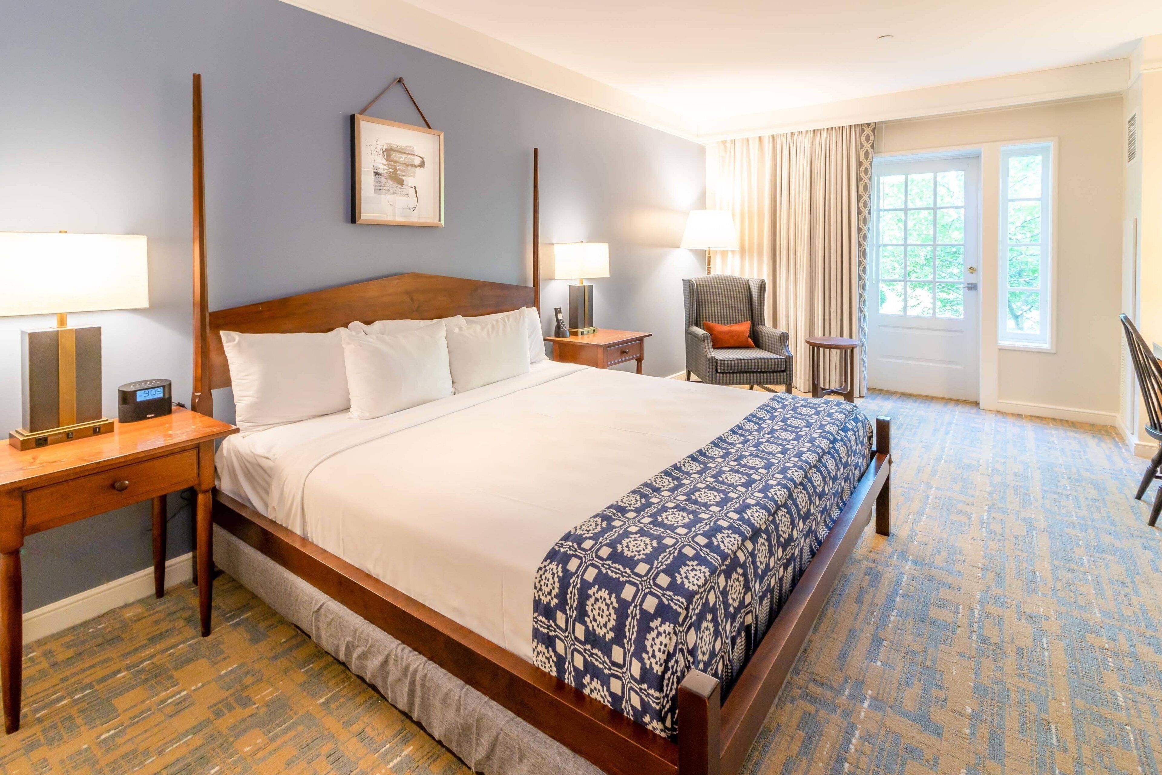 Habitación de hotel con una cama grande, dos mesitas de noche con lámparas, una silla, una puerta con vistas y una alfombra estampada.