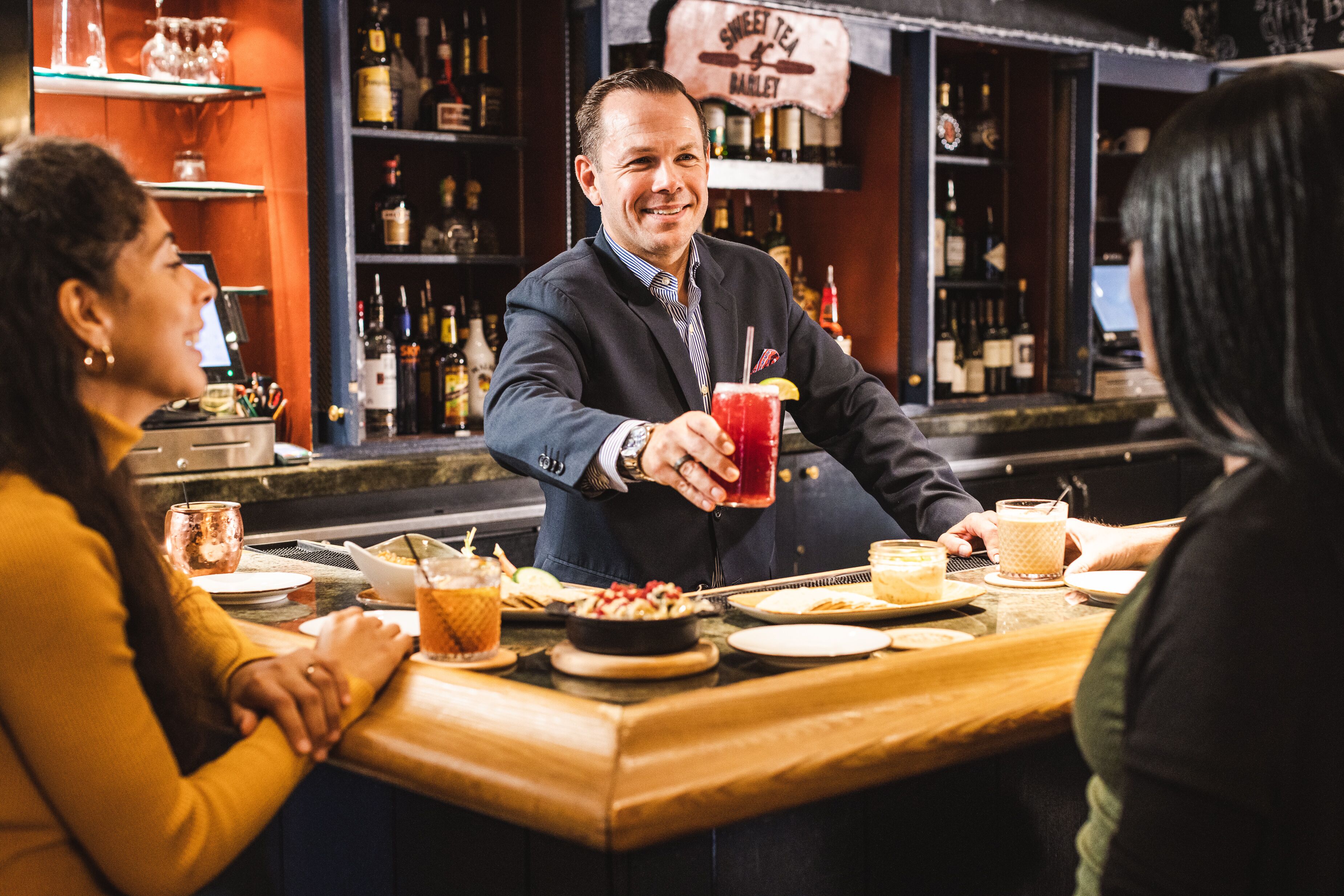 A bar setting with a bartender serving a drink, a woman in a yellow sweater, and a man in a suit.