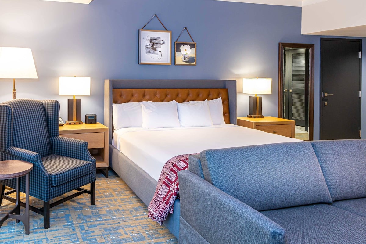 A hotel room with a large bed, a blue armchair, a small round table, and two bedside lamps.