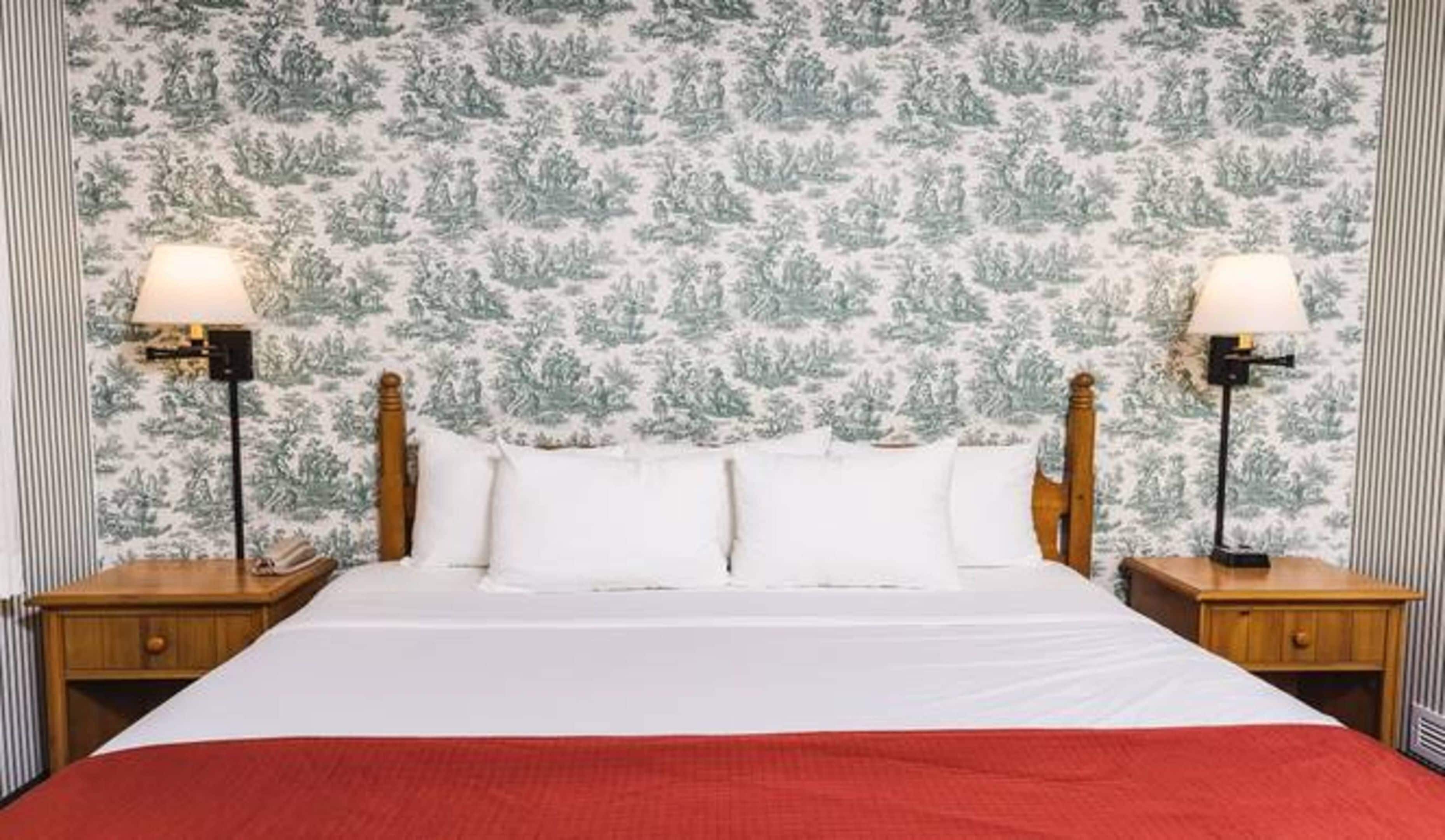 Una cama con sábanas blancas y una manta roja, flanqueada por dos mesitas de noche con lámparas, frente a una pared cubierta de papel tapiz.