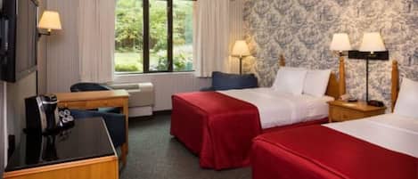 A hotel room with two beds, a desk, a chair, a TV, and a window with a view of greenery.