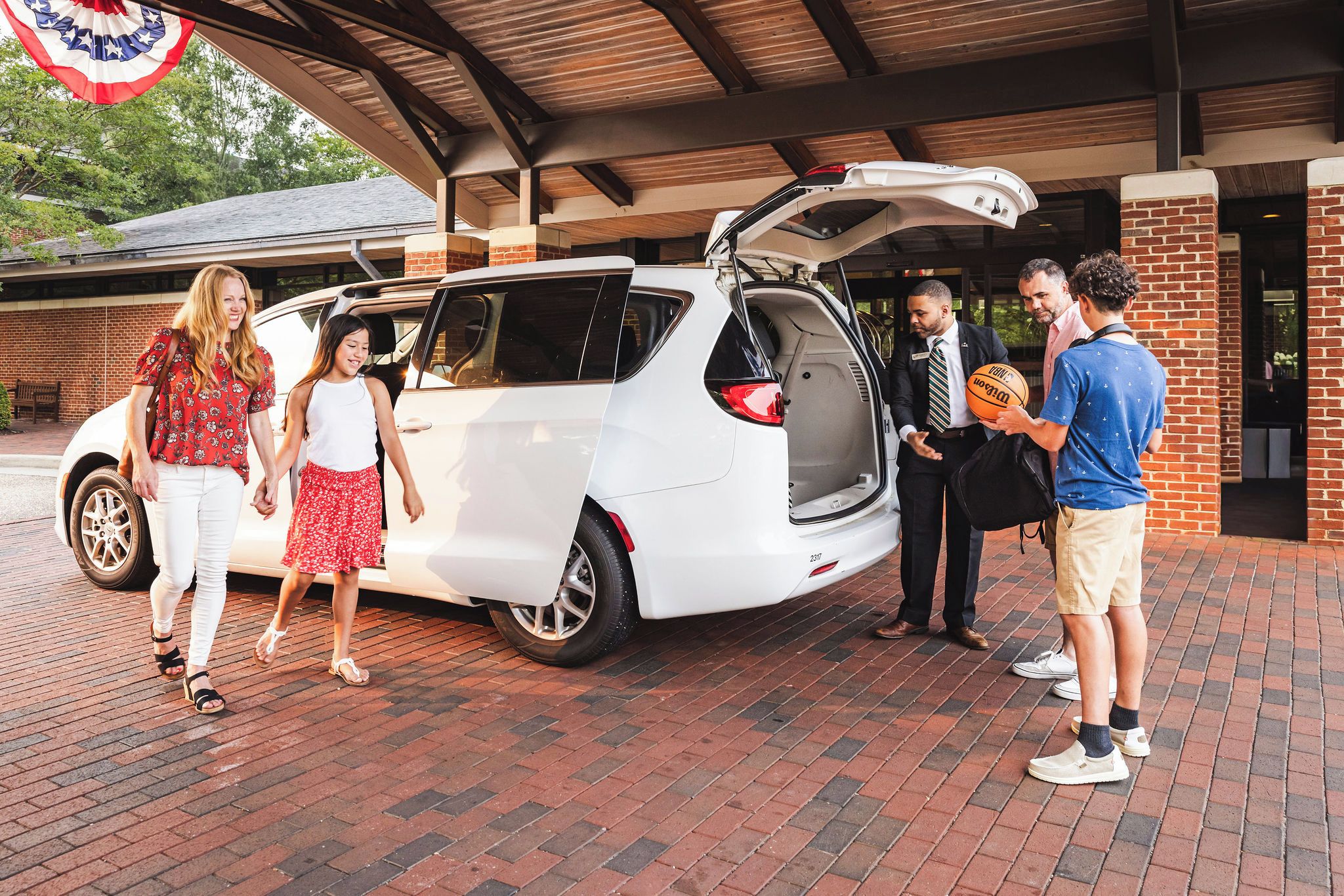 A white minivan parked in a covered parking area with a brick floor, a brick building in the background, and a flag hanging from the ceiling.