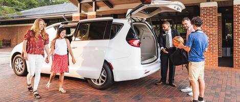A white minivan parked in a covered parking area with a brick floor, a brick building in the background, and a flag hanging from the ceiling.