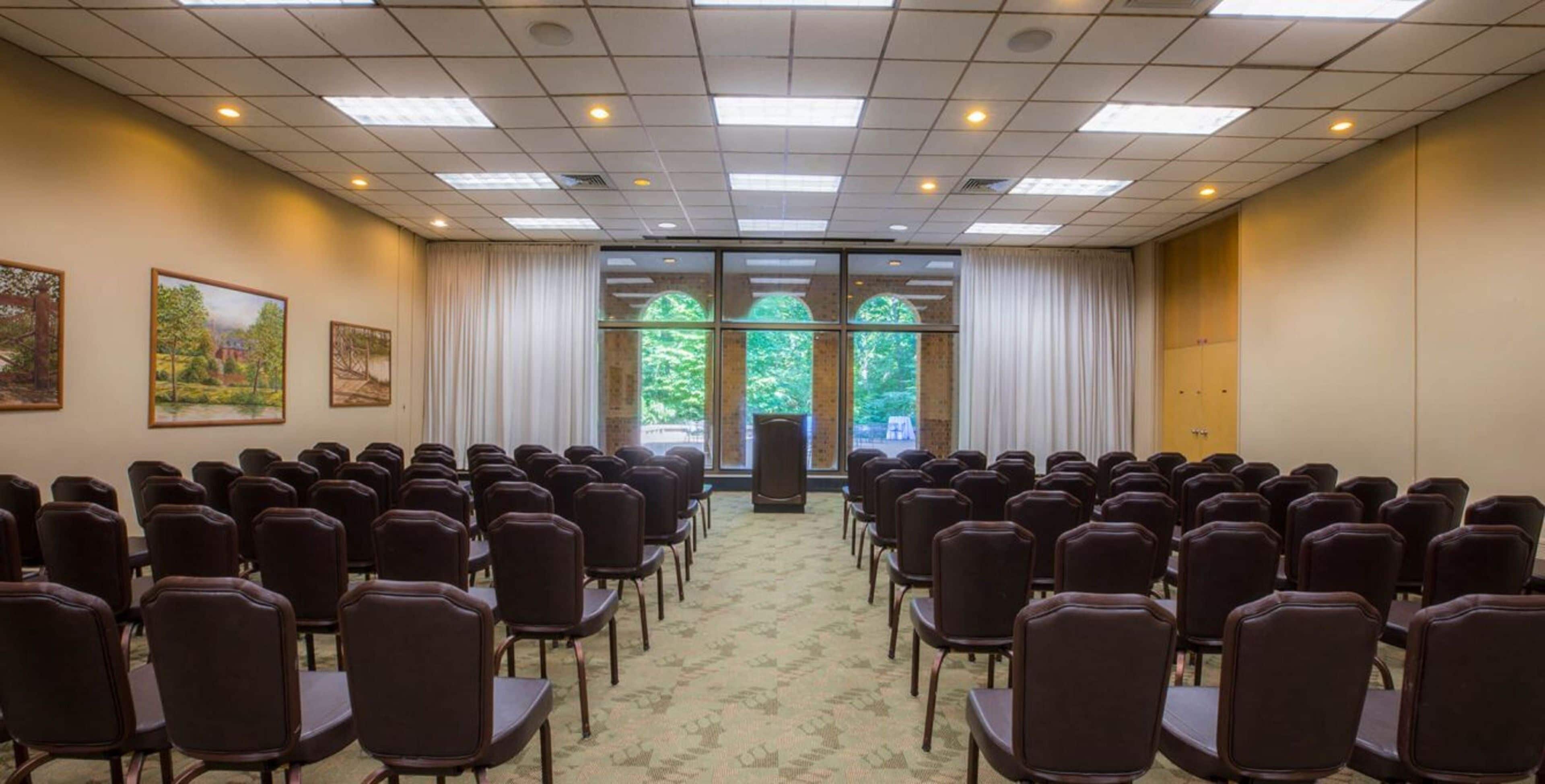 Un salón de conferencias con filas de sillas, amplias ventanas con cortinas y pinturas de paisajes enmarcadas en las paredes.