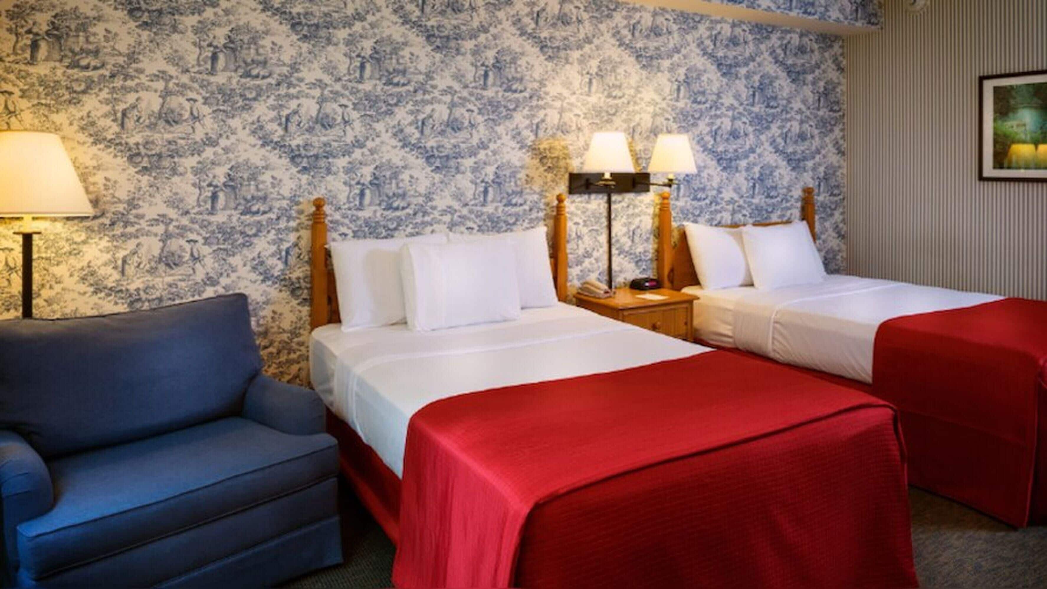 A hotel room with two beds, a blue armchair, and a patterned wallpaper.
