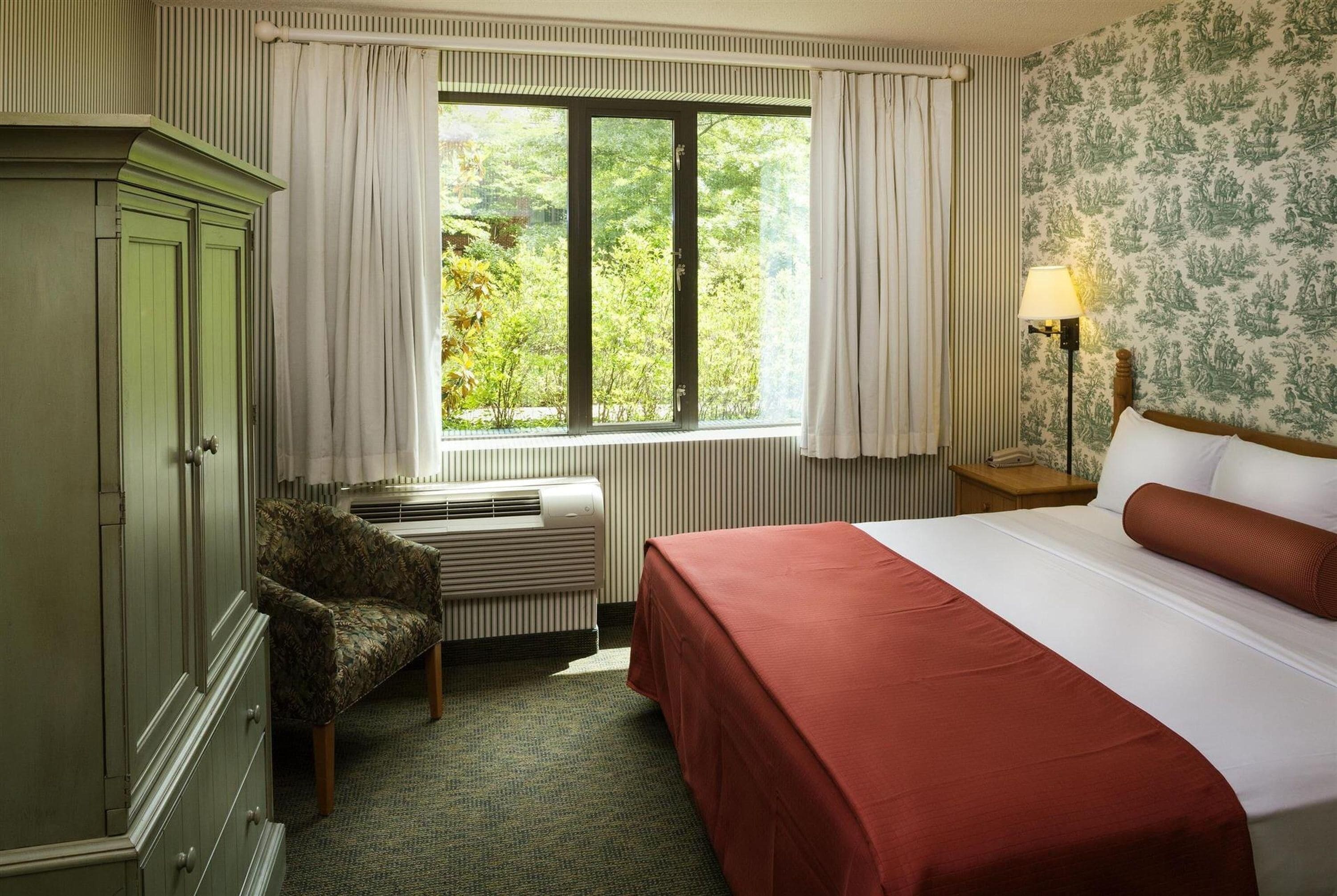 A hotel room with a bed, a wardrobe, a radiator, a window with curtains, and a lamp on a bedside table.