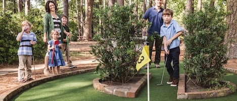 A family at a miniature golf course with a putting green, trees, and a flag.