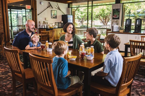 A family dining at a restaurant with a large wooden table, chairs, and a view of the outdoors through large windows.