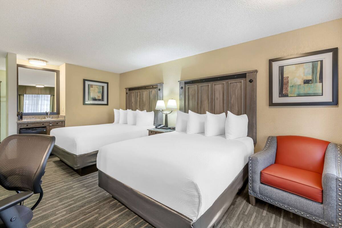 A hotel room with two beds, a desk, a chair, and a lamp.
