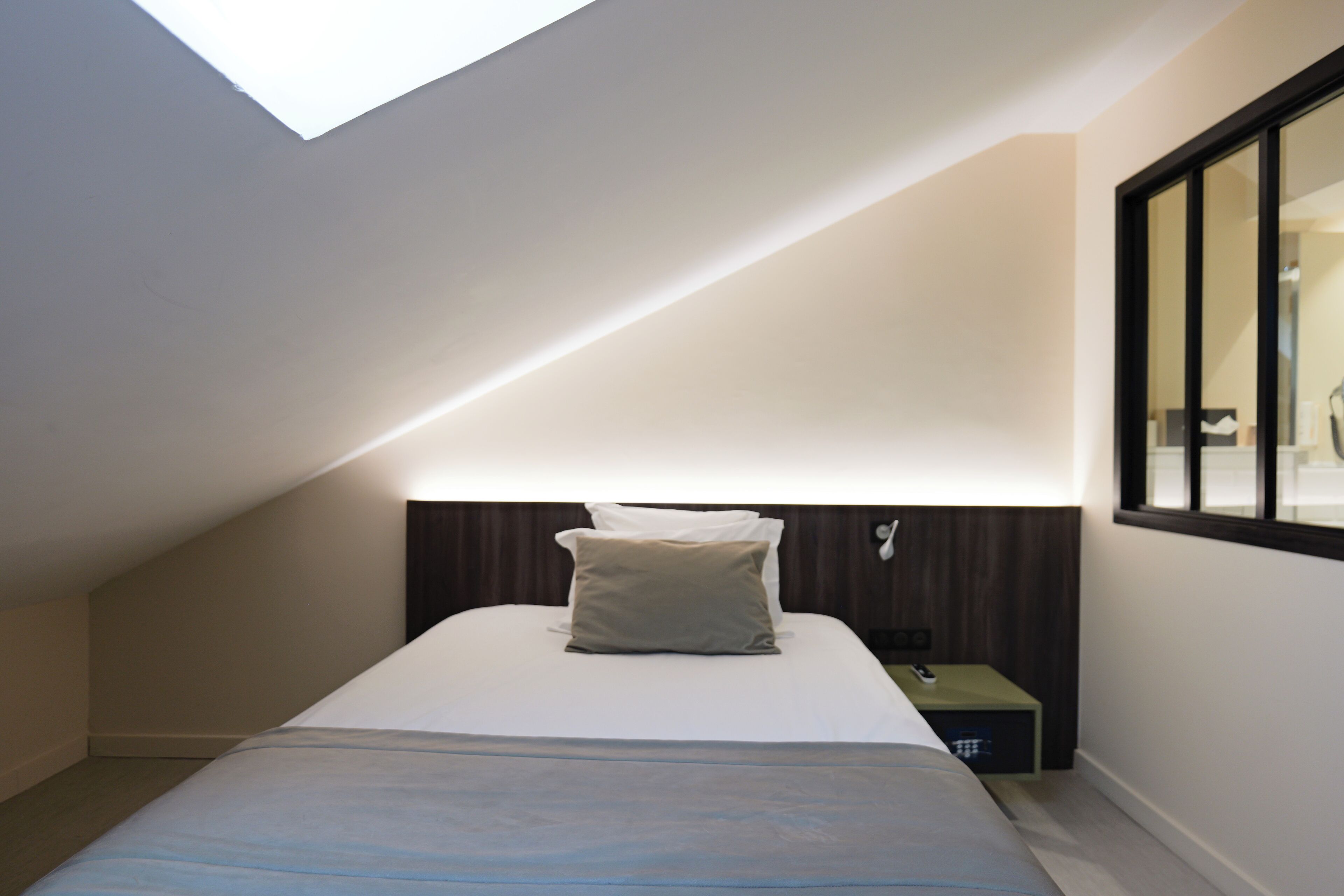 Un dormitorio moderno con tragaluz, una cama con almohada blanca y beige, y una mesita de noche con una caja verde.