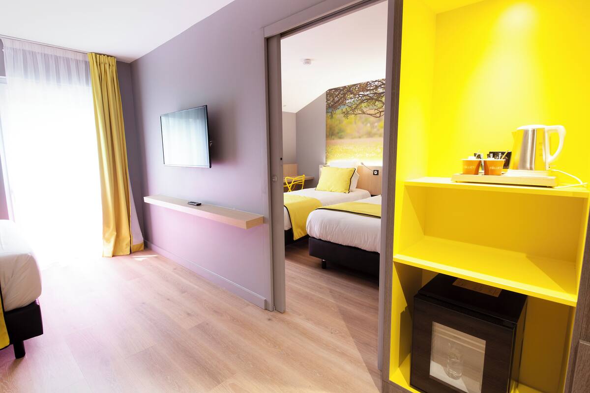 A modern hotel room with a bed, a TV, a small desk, and a bright yellow accent wall.