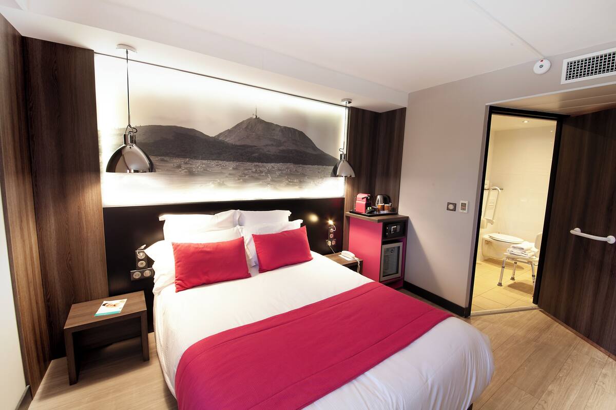A hotel room with a bed, nightstand, lamp, and a view of a mountain through a window.