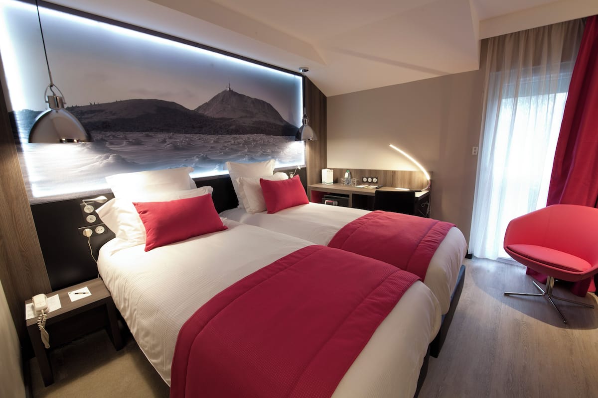 A hotel room with two beds, a desk, a chair, and a large wall-mounted TV displaying a mountain landscape.