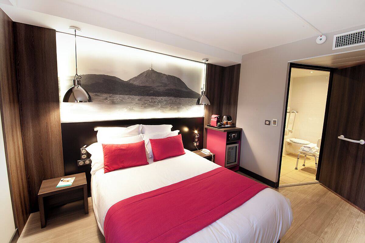 Hotelkamer met een groot bed, rode kussens, een nachtkastje, een bureau, een bureaulamp, een aan de muur bevestigde televisie, en een badkamer zichtbaar door een geopende deur.