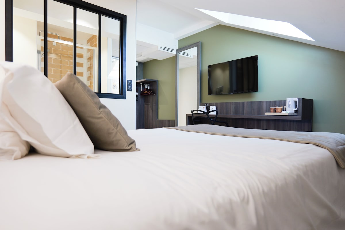 A hotel room with a large bed, a flat-screen TV mounted on the wall, a coffee machine, and a view of the outside through a window.