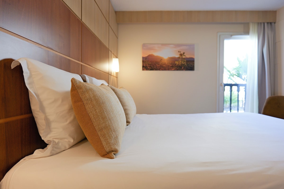 Una cama bien tendida con almohadas blancas y beige, un cabecero de madera y un cuadro de un atardecer en la pared.