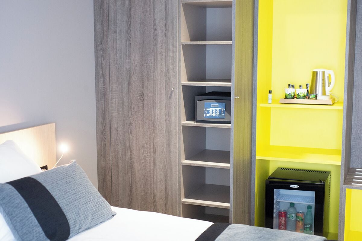 A hotel room with a bed, a grey and black pillow, a wooden wardrobe, a yellow shelf with toiletries, and a mini fridge.